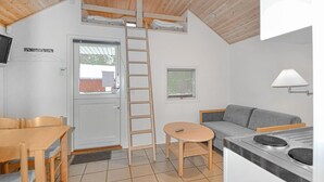 Een kleine keuken met ingebouwde kookplaat, een grijze bank, een eettafeltje en een houten stoel. Een ladder leunt tegen de muur en er is een raam met zonwering te zien.