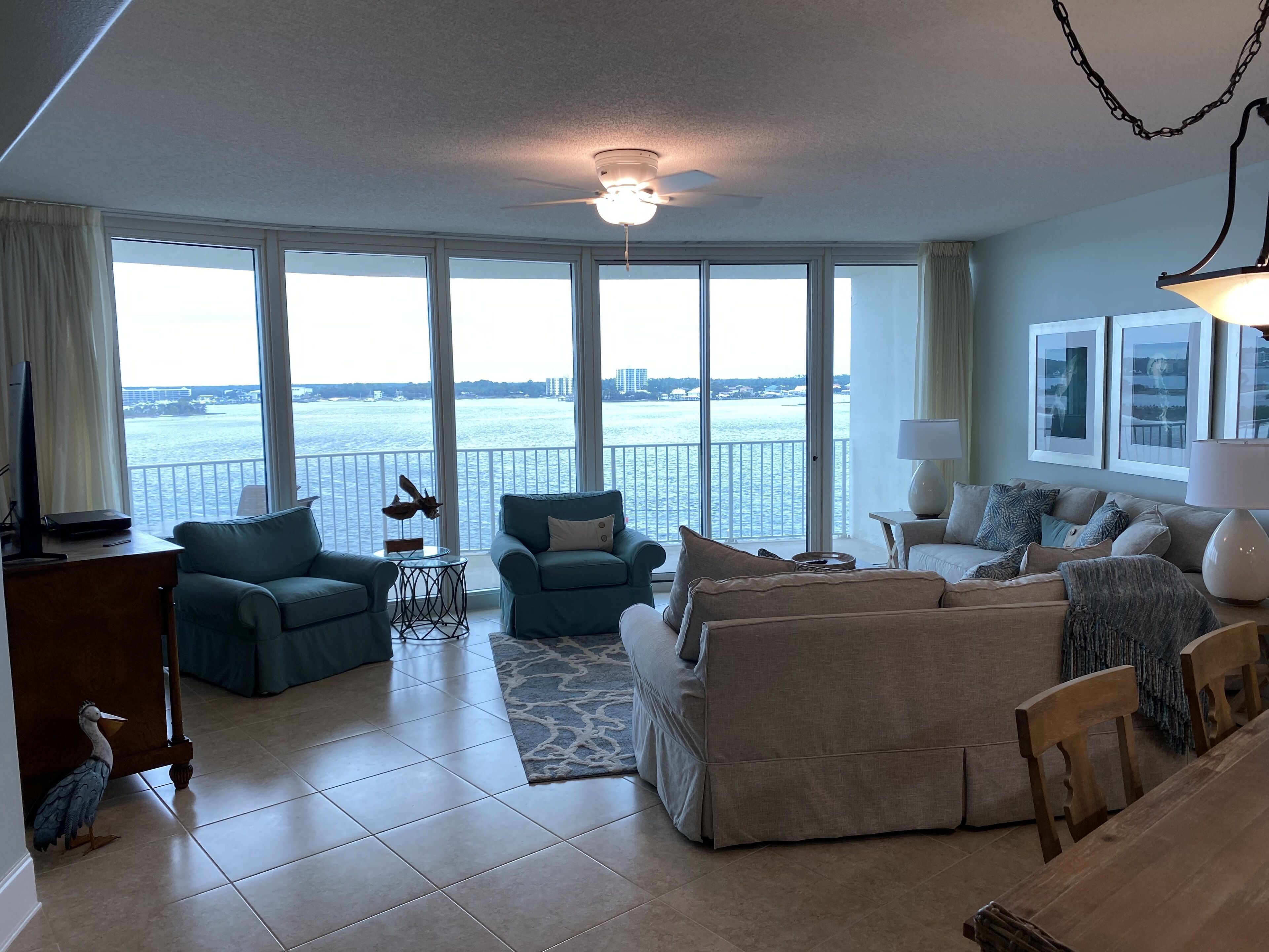 A spacious living room with a large window overlooking a body of water, a ceiling fan, and a dining area with a wooden table and chairs.