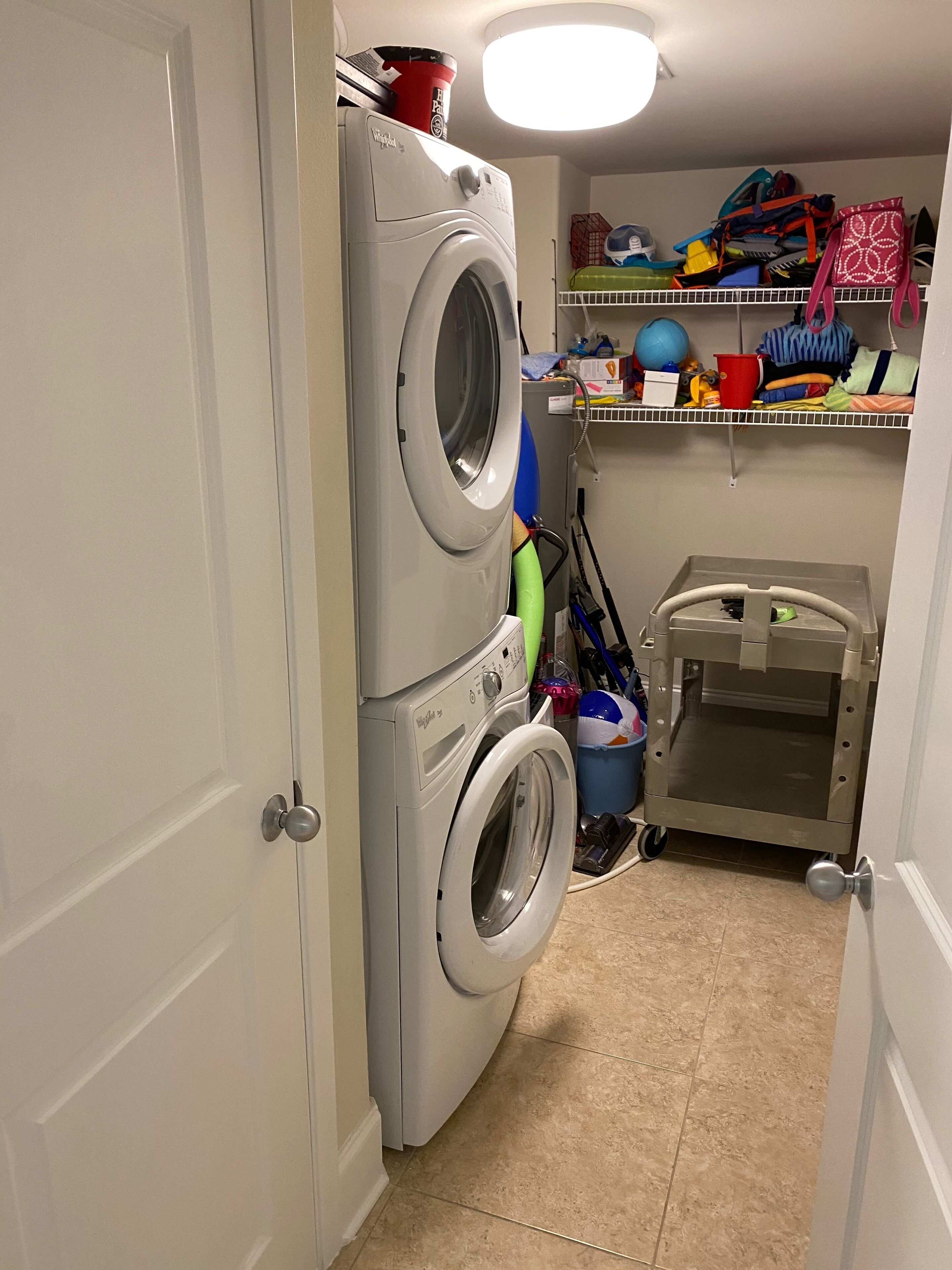 A laundry room with a stacked washer and dryer, a shelf with various items, and a small cart.