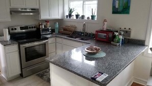 A kitchen with white cabinets, a stainless steel range, a microwave, and a sink. There is a window with plants on the windowsill and a painting on the wall. - Quaint Abingdon Cottage Downtown 420 East Valley Street  (Abingdon)