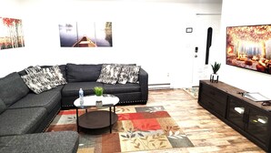 A living room with a dark grey sectional sofa, a round coffee table, and a patterned rug. There are wall-mounted artworks and a television on the wall. - Private Canoes & Charming Lake House  (Long Pond)