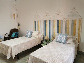 A room with two beds, each with a patterned bedspread and matching pillows. There is a wooden chair and a small table with a plant and a lamp. The wall features a striped panel design and a ceiling light fixture.