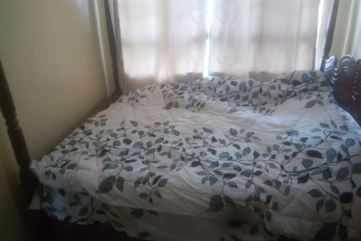 A bed with a floral bedspread, a wooden headboard, and a window with sheer curtains.