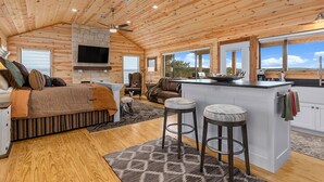 A spacious cabin interior with a large living area, a kitchen with a central island, and a cozy seating area. - No Rancho Properties Texas Haus | Hot Tub | Beautiful Views (Fredericksburg)