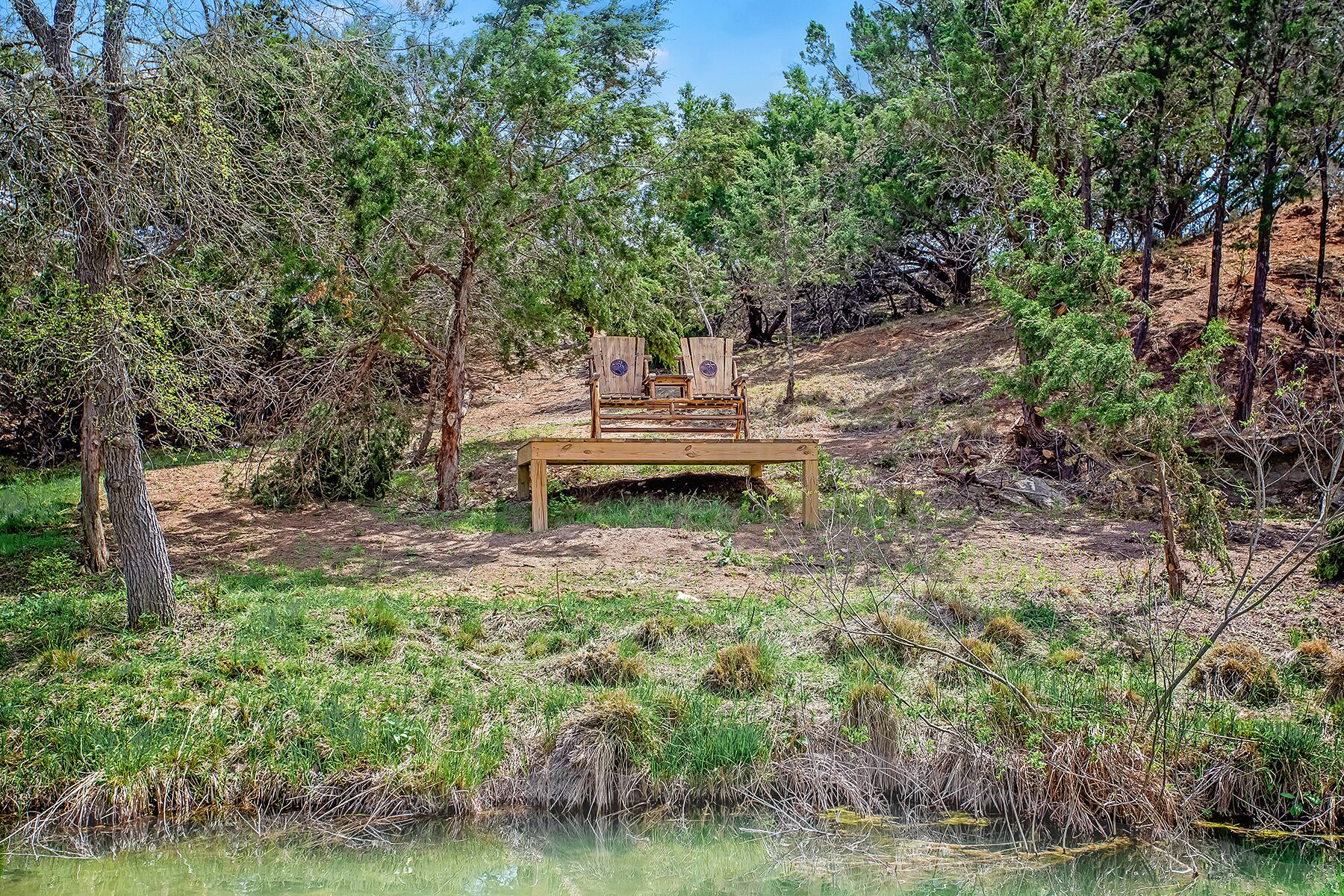 Un banco de madera con dos sillas en una zona boscosa cerca de un cuerpo de agua.