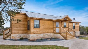 A wooden cabin with a stone foundation, a metal roof, and a small porch with a staircase. - No Rancho Properties Texas Haus | Hot Tub | Beautiful Views (Fredericksburg)