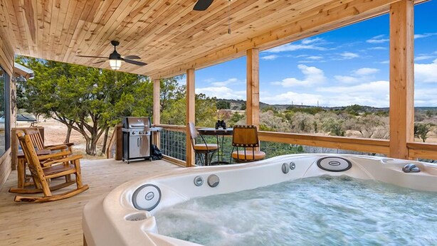 A wooden deck with a hot tub, seating, and a grill. - No Rancho Properties Texas Haus | Hot Tub | Beautiful Views (Fredericksburg)