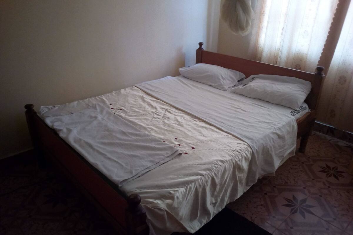 Ein Einzelbett mit hölzernem Gestell und weißer Bettwäsche in einem Zimmer mit gestreiften Bodenfliesen.