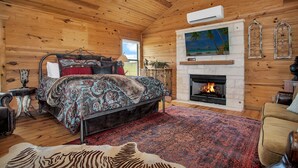 Une chambre située dans une cabane en rondins, avec un lit, une cheminée, un ventilateur de plafond et une grande fenêtre.