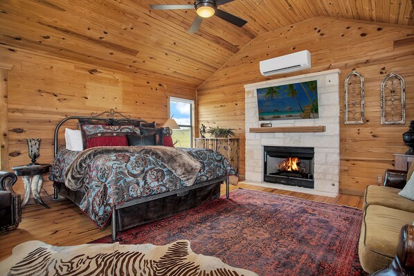 Ein Schlafzimmer in einer Blockhütte mit Bett, Kamin, Deckenventilator und großem Fenster.