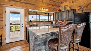 Une cuisine rustique aux murs en bois, avec un grand îlot central doté de tabourets et une vue sur l’extérieur par la porte.