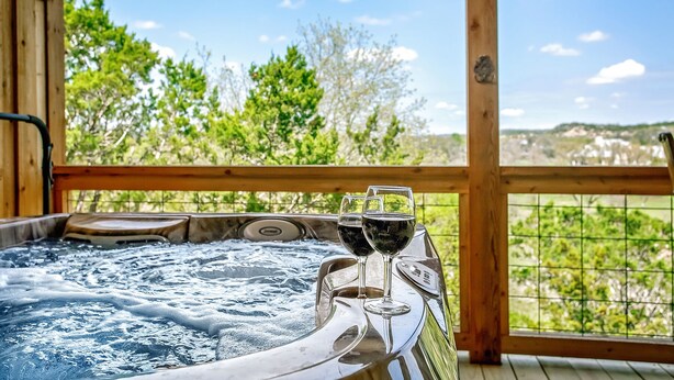 Un bain à remous avec deux verres de vin, installé sur une terrasse en bois offrant une vue sur la verdure et les collines.