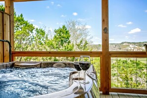 A hot tub with two glasses of wine on a wooden deck with a view of greenery and hills.
