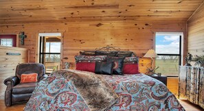Ein gemütliches Schlafzimmer in einer Hütte mit Holzbalkendecke, einem Bett mit gemusterten Textilien, einem Ledersessel und Blick ins Freie.