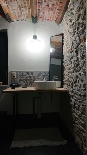 Ein Badezimmer mit Steinwand, gefliestem Rücksprüher, einem runden Waschbecken, einem Spiegel und einer Deckenleuchte.