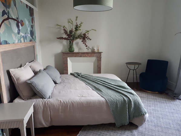 A bedroom with a bed, a nightstand, a chair, a fireplace, and a painting on the wall.