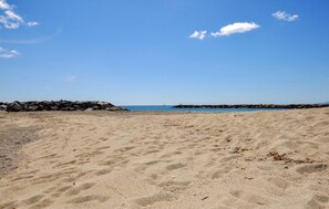 Ein sandiger Strand mit einem Felsenmole im Hintergrund und blauem, klarem Himmel.