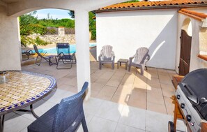 Eine Terrasse im Freien mit einem gefliesten Tisch, Stühlen, einem Grill und Blick auf einen Swimmingpool.