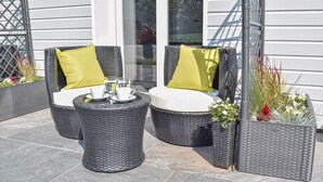 A patio with two wicker chairs and a small table, each with a cushion. There are potted plants and a glass door in the background.