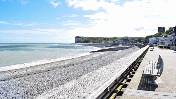 A pebbled beach with a promenade, a cliff, and buildings in the background.