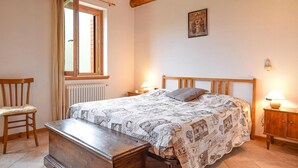A bedroom with a wooden bed, a nightstand with a lamp, a chair, a wooden bench, a window with curtains, and a radiator. - 3 bedroom beautiful home in Sovramonte (Sovramonte)