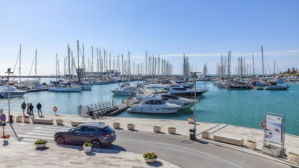 A marina with numerous sailboats docked, a pedestrian walkway, and a parked car.