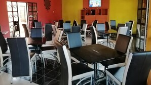 Un comedor con varias mesas y sillas, un televisor montado en la pared y un ventanal con reja decorativa.
