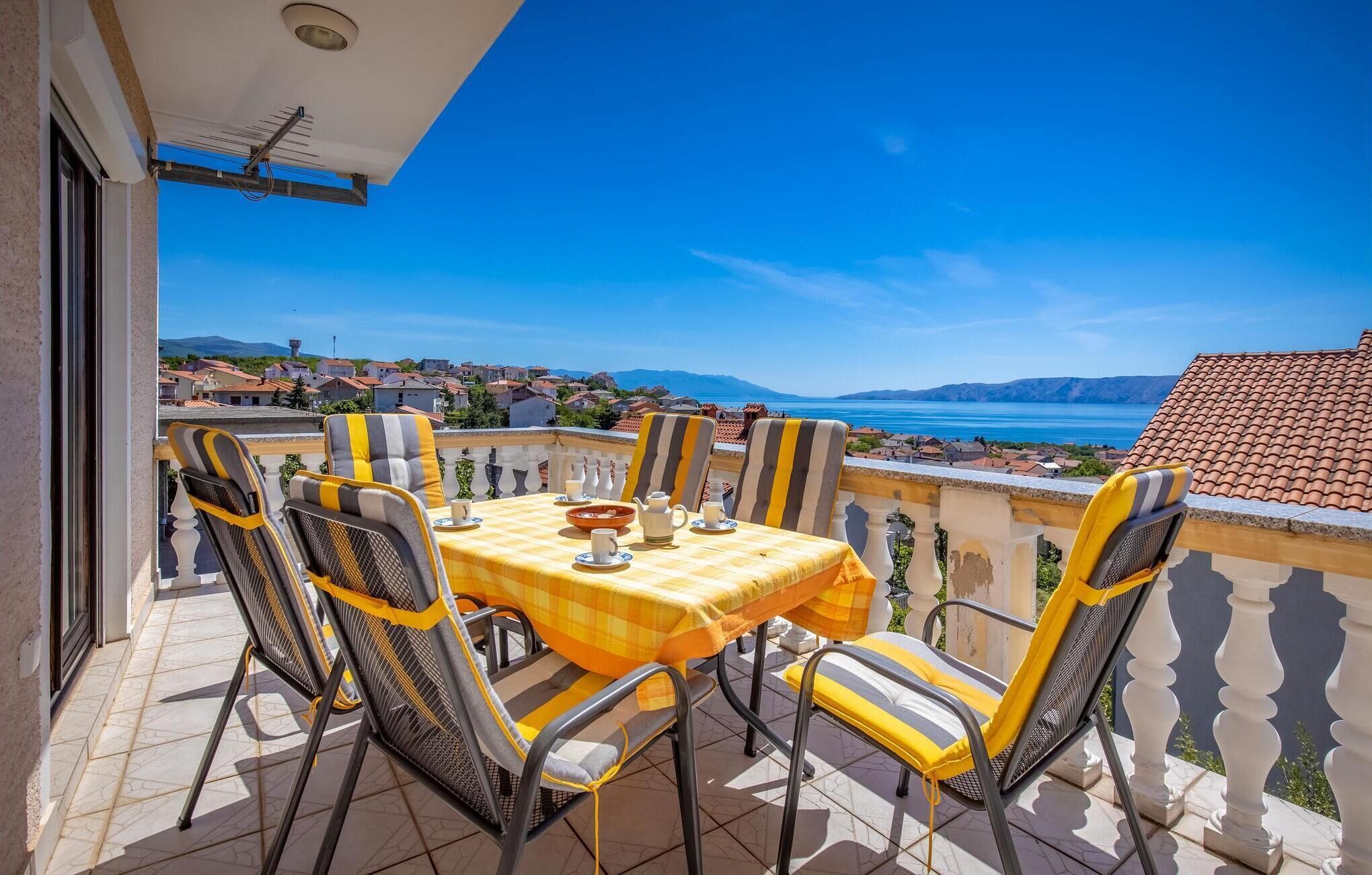 A balcony with a checkered tablecloth, striped chairs, and a view of a coastal town and sea.
