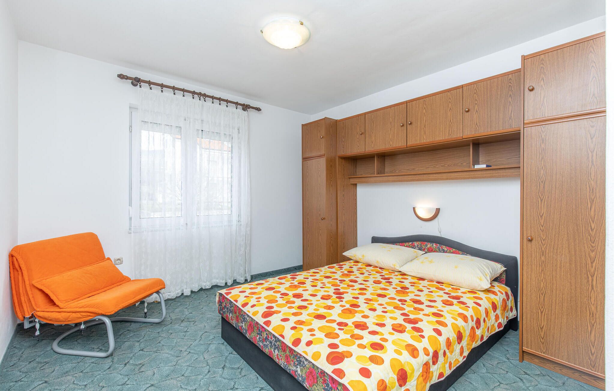 A bedroom with a bed, an orange chair, wooden wardrobe, and a window with curtains.