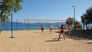Un match de volleyball de plage se déroule sur un terrain sablonneux, avec un filet, des arbres et une vue dégagée sur la mer.