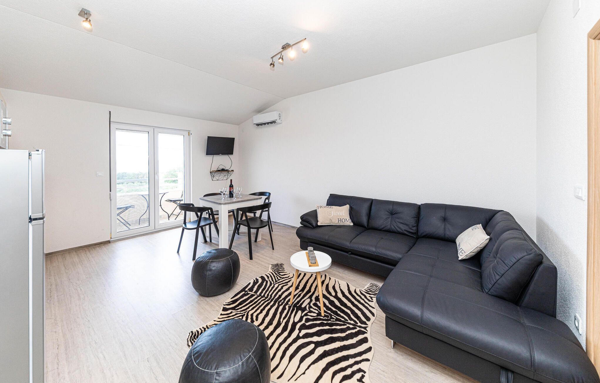 Un salon moderne avec un canapé en cuir noir, un tapis à motifs zébrés, un pouf rond, une petite table blanche et un coin repas comprenant une table blanche et des chaises noires.