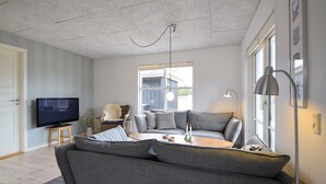 Uma sala de estar moderna com um sofá cinzento, uma televisão, uma cadeira de vime e uma janela ampla.