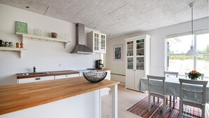 Uma cozinha moderna com armários brancos, bancada de madeira e uma tigela de arame preta. Há uma área de jantar com mesa e cadeiras, e uma vista do exterior através de grandes janelas.