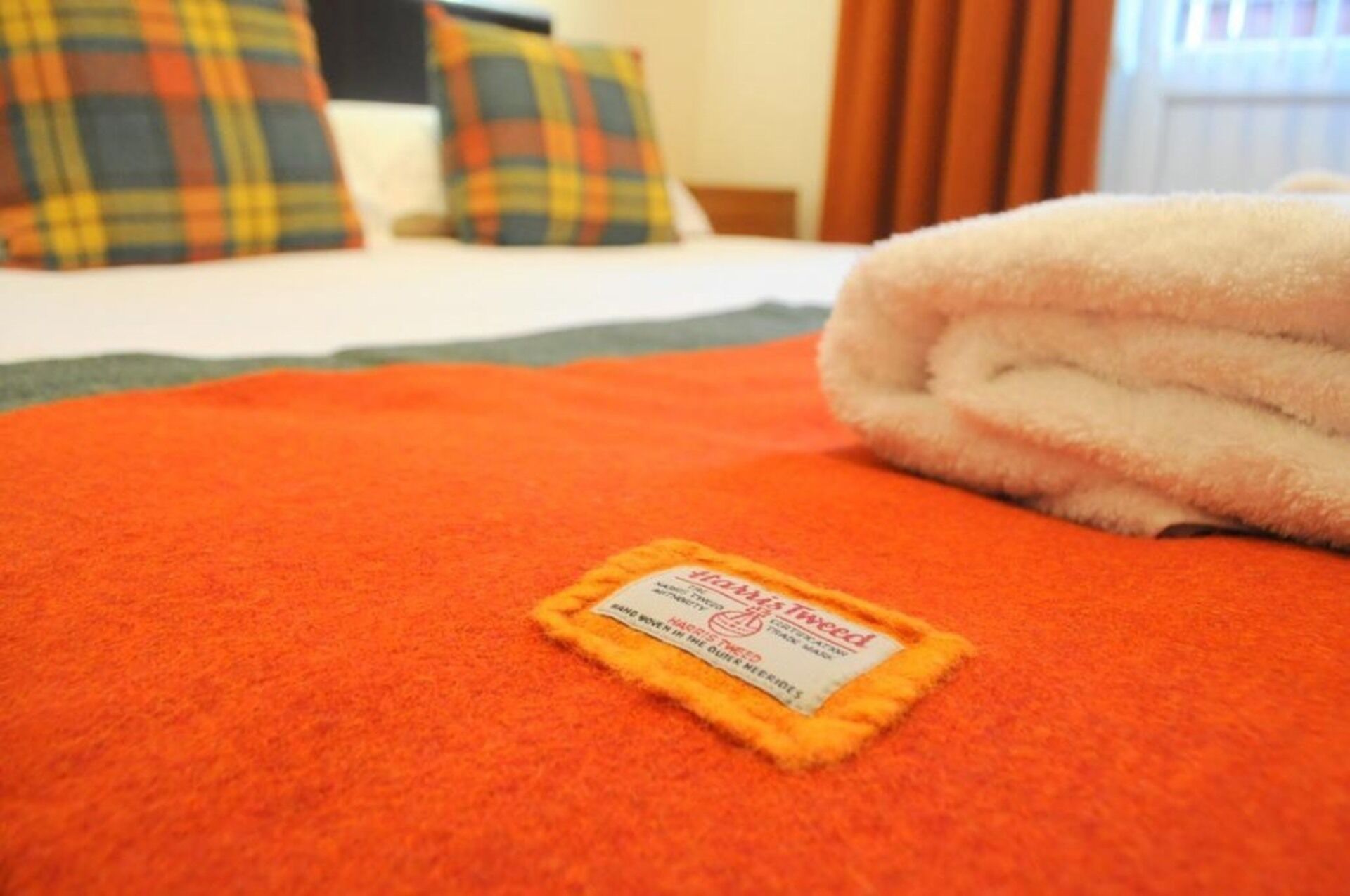 Cama bem arrumada com uma toalha branca dobrada e um cobertor laranja. Uma etiqueta no cobertor diz 'Tema Hogwarts'.