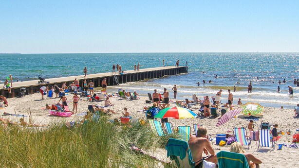 En strand med en mole, strandstole, parasoller og mennesker, der nyder havets nærhed.