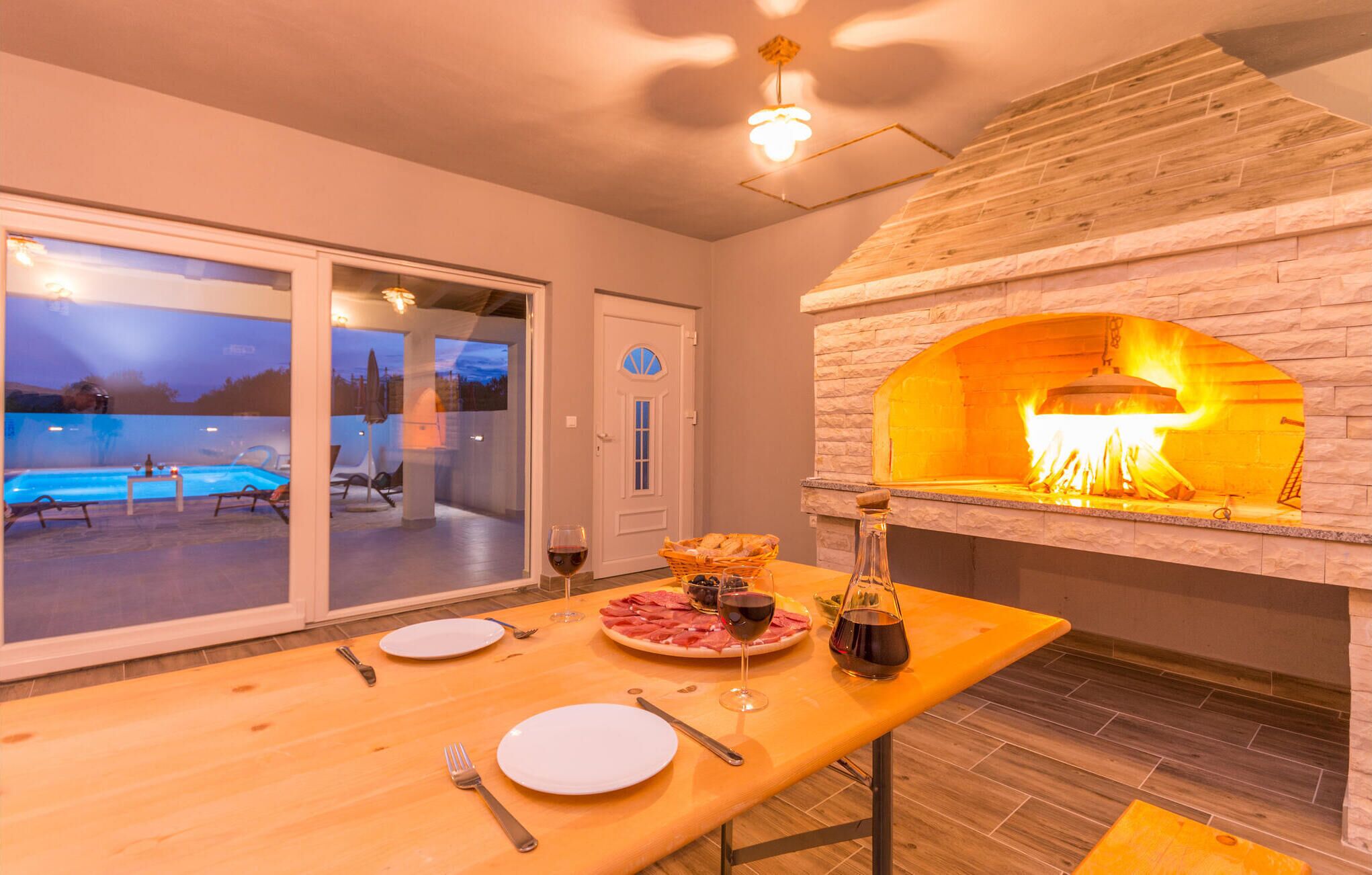 Comedor con chimenea de piedra, piscina visible a través de puertas de vidrio y mesa preparada para dos con platos, cubiertos y una botella de vino.