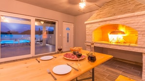 A dining area with a stone fireplace, a pool visible through glass doors, and a table set for two with plates, cutlery, and a bottle of wine. - Stunning home in Benkovac (Benkovac)