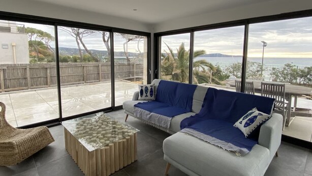 A modern living room with a large glass sliding door leading to a patio, a sofa, a coffee table, and a view of the sea. - Villa Récampado - Feet in the water, pool and sea (Cavalaire-sur-Mer)