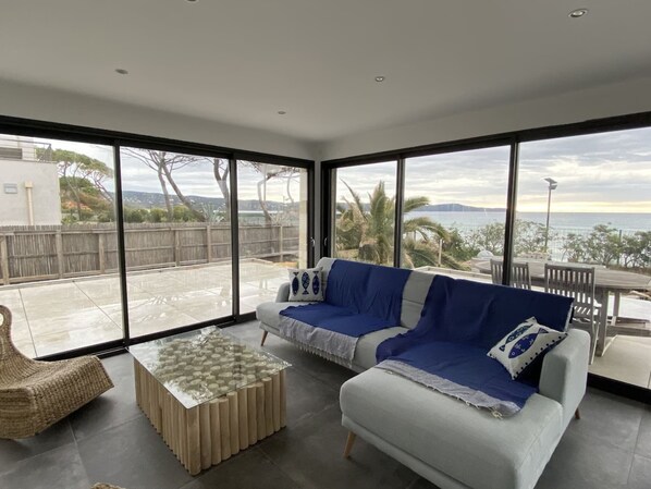 Een moderne woonkamer met een grote glazen schuifdeur naar een terras, een bank, een salontafel en uitzicht op zee.