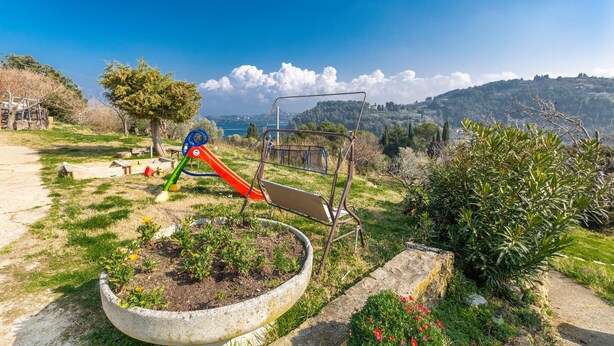 Ein Spielplatz mit Rutsche und Schaukeln, umgeben von einem Garten mit einem großen Pflanzkübel und blühenden Pflanzen.