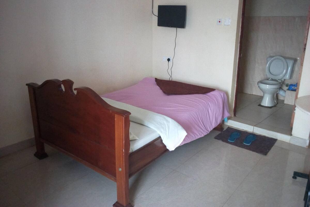 A small, simple room with a wooden bed, a wall-mounted TV, and a toilet in the corner.