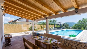 Un espacio al aire libre techado con piscina, mobiliario de madera y vistas al paisaje circundante.