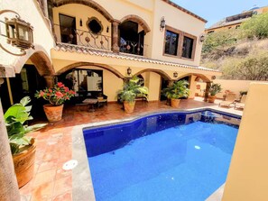 Área de piscina com terraço, plantas em vasos e um edifício com arcos e varanda.