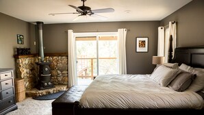 A bedroom with a bed, a stone fireplace, a ceiling fan, and a view of the outdoors through a glass door. - Gorgeous Riverfront Home, VHR# 073569, TOT# T65183 (Lotus)