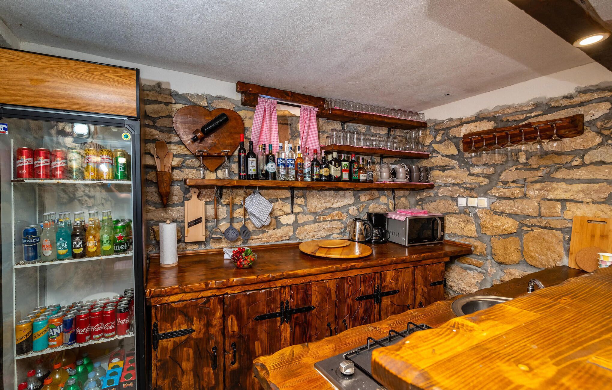Un espace bar avec un mur en pierre, des étagères en bois remplies de bouteilles, un réfrigérateur contenant des boissons et un comptoir en bois avec un évier.