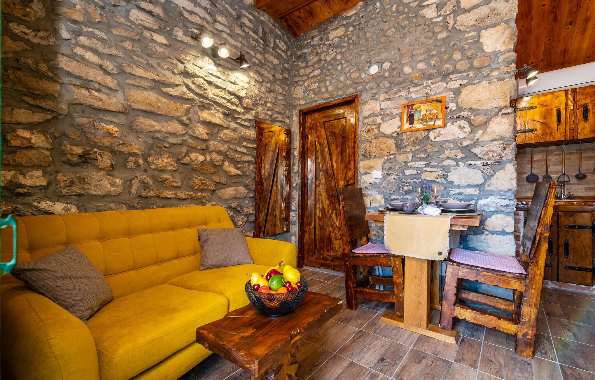 Un interior rústico con pared de piedra, muebles de madera, un sofá amarillo, un frutero y una zona de cocina con armarios de madera y un lavamanos.
