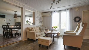 Ein Wohnzimmer mit Sofa, Ohrensessel, Hocker und Couchtisch. Es gibt eine Essecke mit Tisch und Stühlen. Der Raum hat Holzdielen und weiße Wände mit Wandverkleidung.