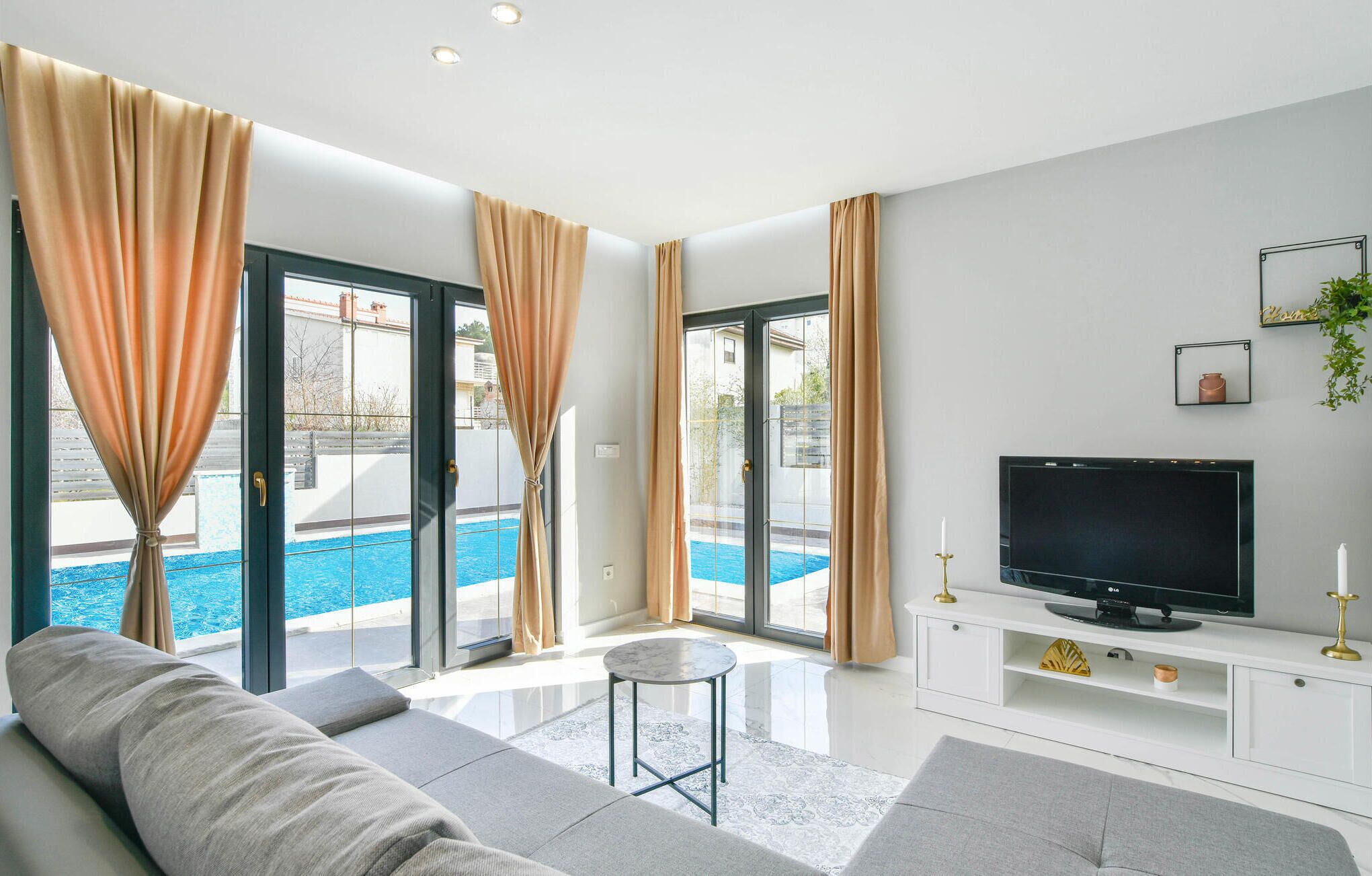 A modern living room with a sofa, a TV on a white entertainment unit, and a pool visible through the glass doors.