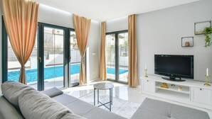 A modern living room with a sofa, a TV on a white entertainment unit, and a pool visible through the glass doors. - Luxurious home in Split (Split)