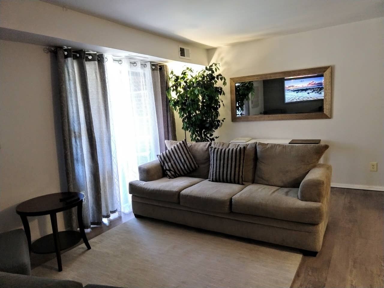 A living room with a beige sofa, two striped pillows, a small round table, a potted plant, and a TV mounted on the wall.
